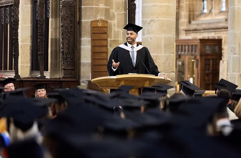 Leon Greenwood in academic gown and cap, speaking from a lectern to graduates in a cathedral
