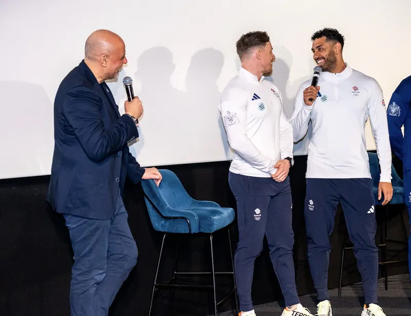 Leon Greenwood speaking into a microphone on stage beside a Team GB teammate and an event host